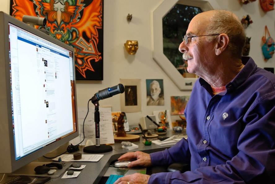 Sociologist and author Howard Rheingold at his home office, sitting at his desk in front of a large computer monitor, holding a computer mouse in his right hand.
