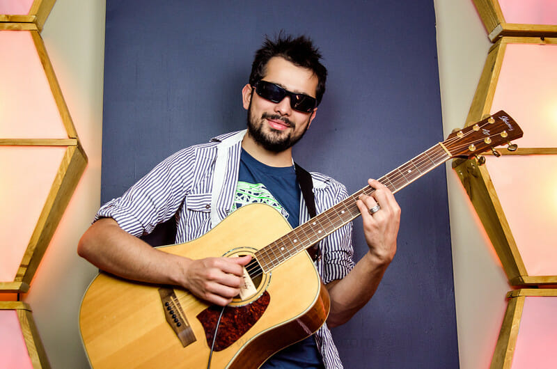 Portrait of Youtube star Joe Pena, known as MysteryGuitarMan, wearing sunglasses and holding a guitar, in front of a colorful studio background.