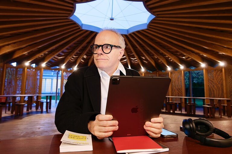 Hans Ulrich Obrist, Artistic Director at the Serpentine Galleries in London, sits in the wooden pavilion designed by architect Lina Ghotmeh (2023), holding an iPad, looking to the left.